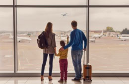 Ein Mann, eine Frau und ein Kind im Grundschulalter stehen mit ihrem Handgepäck an einer großen Scheibe enes Flughafens und betrachten die abhebenden Flugzeuge.