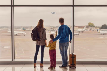 Ein Mann, eine Frau und ein Kind im Grundschulalter stehen mit ihrem Handgepäck an einer großen Scheibe enes Flughafens und betrachten die abhebenden Flugzeuge.