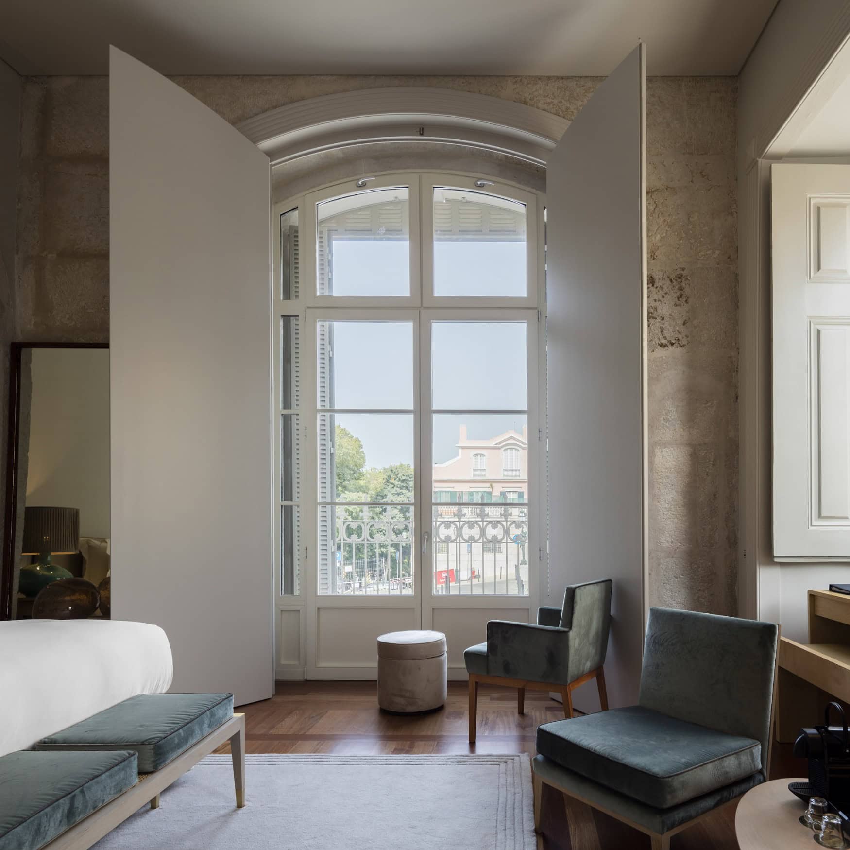 Ausschnitt aus einem Hotelzimmer im Verride Palácio in Lissabon; zentral ein hohes Doppelfenster, links ein freistehendes Bett, rechts ein flacher, blauer Polstersessel.