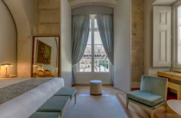 Ausschnitt aus einem Hotelzimmer im Verride Palácio in Lissabon; zentral ein hohes Doppelfenster, links ein freistehendes Bett, rechts ein flacher, blauer Polstersessel.