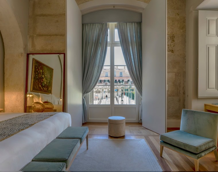 Ausschnitt aus einem Hotelzimmer im Verride Palácio in Lissabon; zentral ein hohes Doppelfenster, links ein freistehendes Bett, rechts ein flacher, blauer Polstersessel.