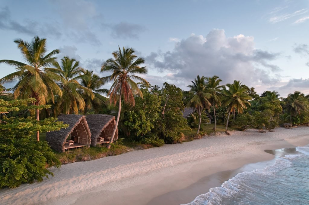 Luftaufnahme des Strands am Voaara auf Madagaskar im Abendlicht: zwei Spa-Pavillons mit gewölbten Strohdächern am Waldrand, davor ein langer weißer Sandstrand mit sanften Wellen.