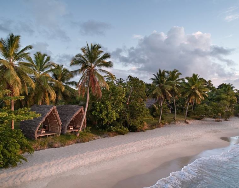Luftaufnahme des Strands am Voaara auf Madagaskar im Abendlicht: zwei Spa-Pavillons mit gewölbten Strohdächern am Waldrand, davor ein langer weißer Sandstrand mit sanften Wellen.