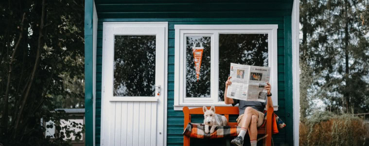 Mann liest Zeitung neben einem West Highland White Terrier auf der Bank einer grün-weißen Anglerhütte im Harz.