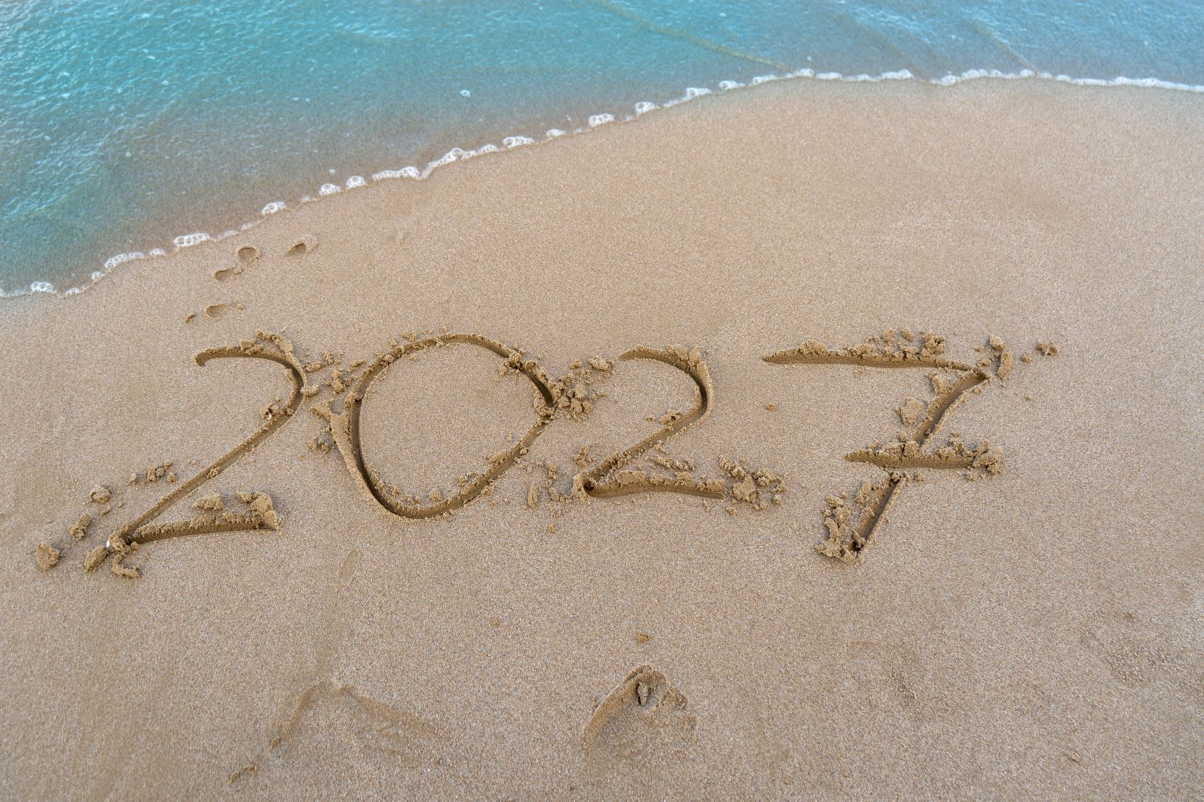 Jahreszahl 2027 im Sand an einem Strand mit Meer und Fußspuren
