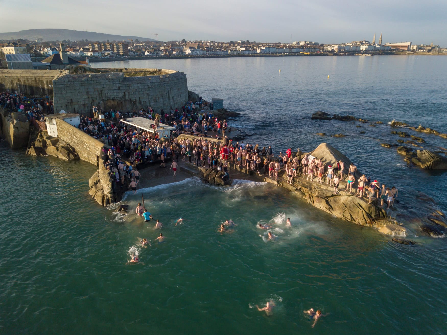 Luftaufnahme des Neujahrsschwimmens am Forty Foot bei Dublin. Zahlreiche Menschen stehen auf den Felsen an der Bucht, einige schwimmen im kalten Meerwasser.