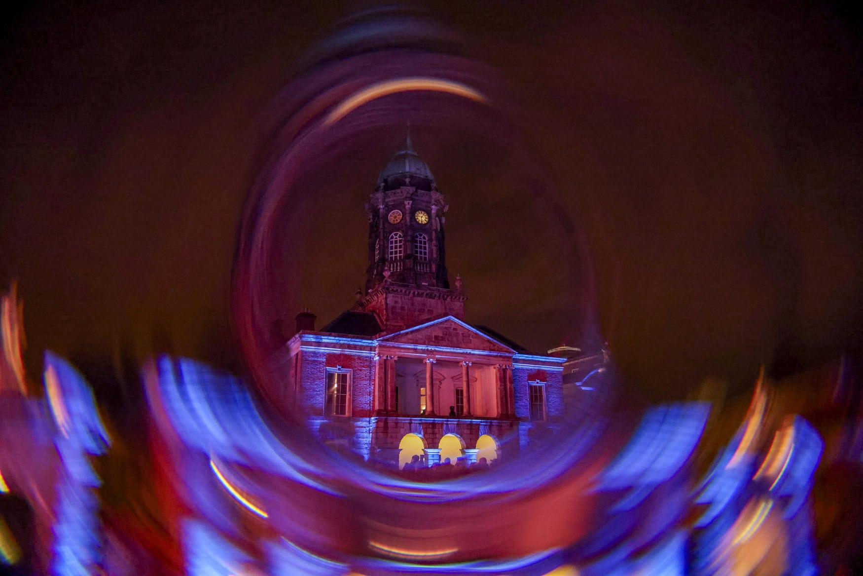 Der beleuchtete Uhrturm von Dublin Castle bei Nacht, aufgenommen durch einen kreisförmigen Lichteffekt aus blauen und roten Streifen. Vor dem Gebäude ist eine Menschenmenge zu erahnen.