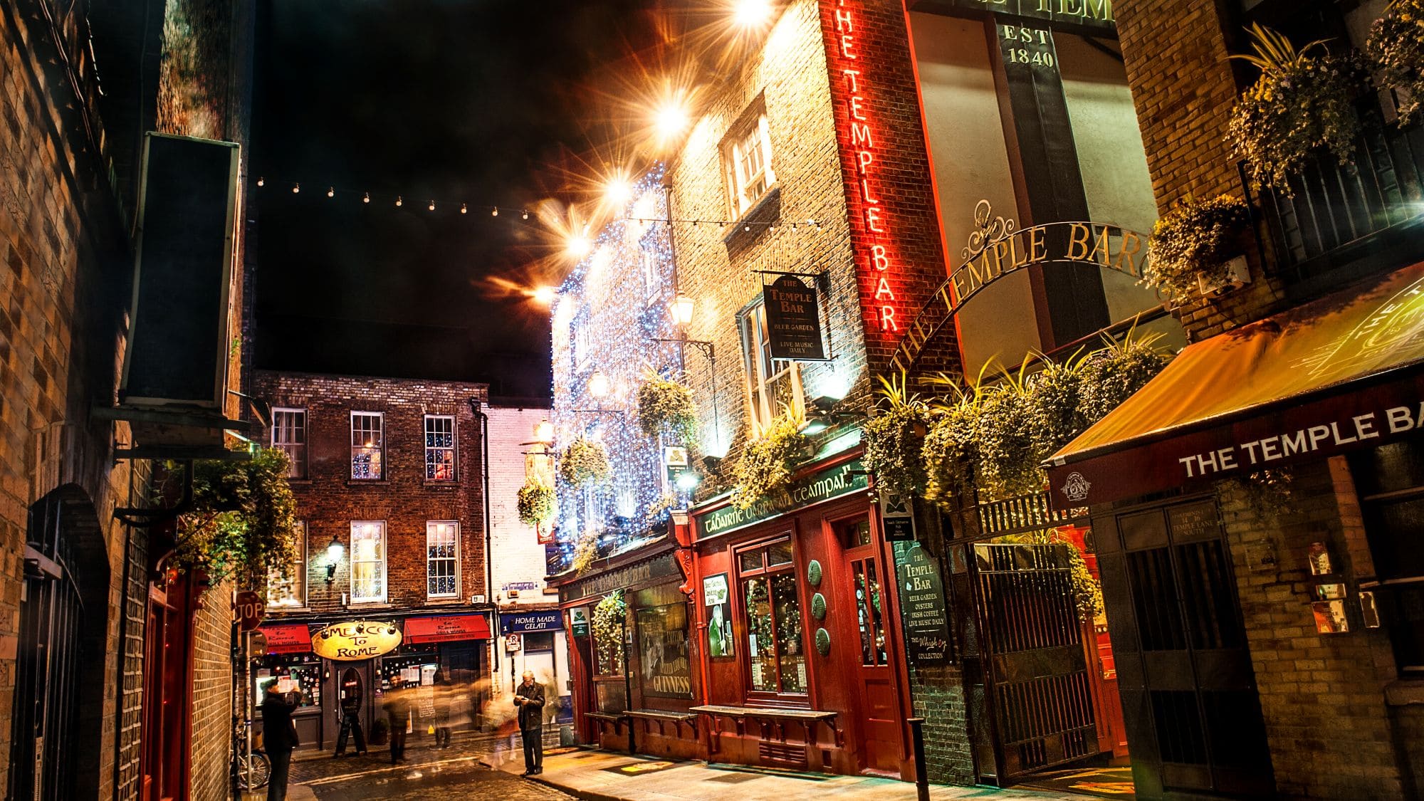 Die beleuchtete Fassade des Pubs The Temple Bar in Dublin bei Nacht. Über der Gasse hängen Lichterketten, dazu schmücken Blumenampeln und weihnachtliche Beleuchtung die Häuserzeile.