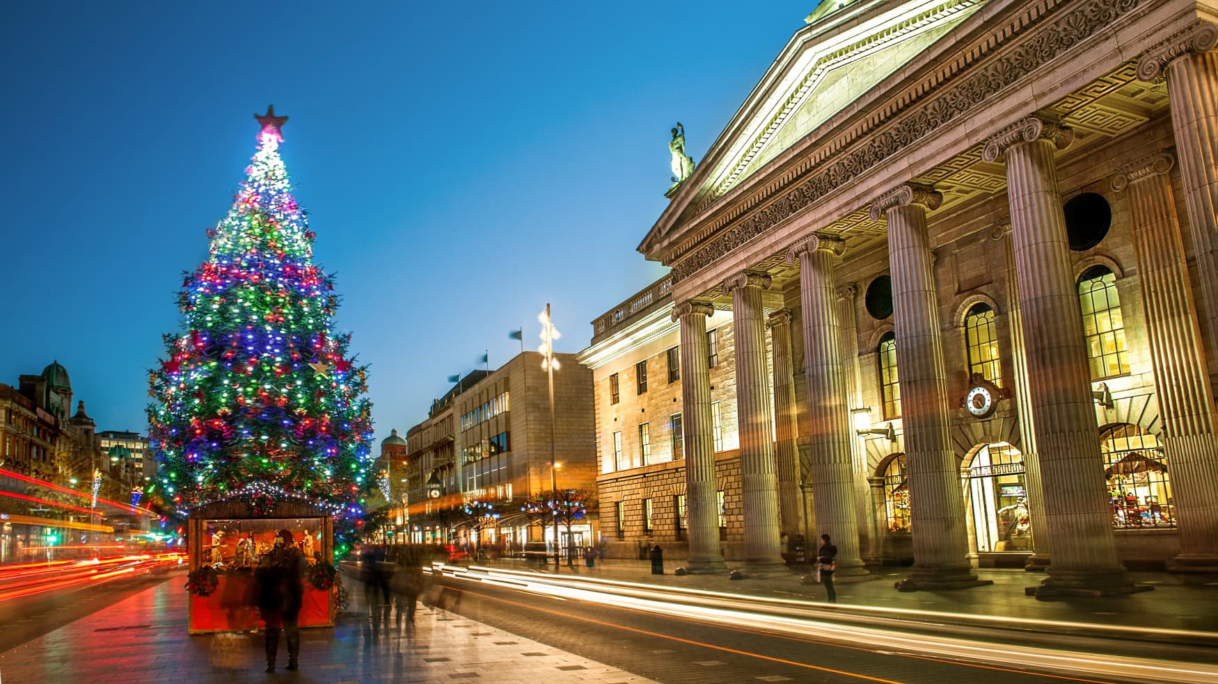 Ein hoch geschmückter Weihnachtsbaum leuchtet abends an der O'Connell Street in Dublin vor dem beleuchteten General Post Office mit seinen Säulen. Lichtspuren des Verkehrs ziehen über die nasse Straße.