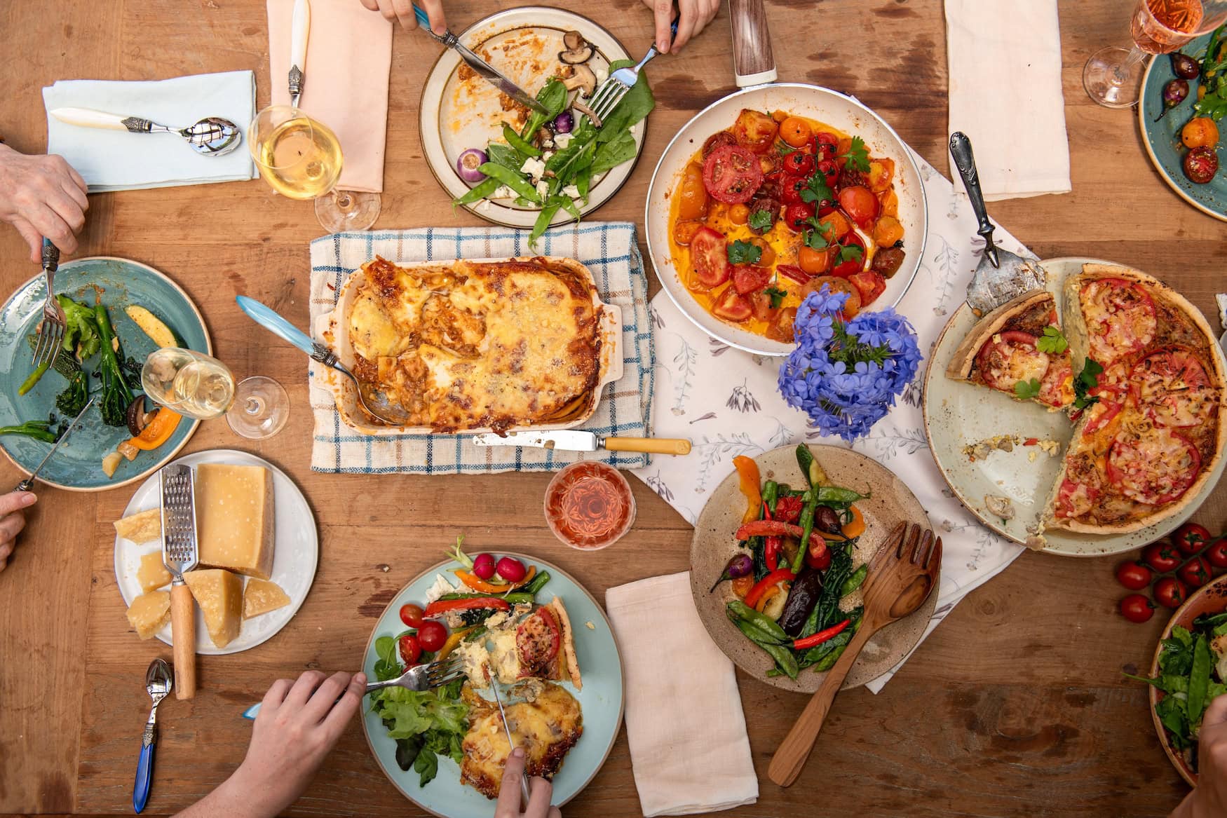 Blick von oben auf einen Holztisch mit italienischer Familienmahlzeit, Lasagne, Tomatentarte, Salaten und Wein, viele Hände greifen zu