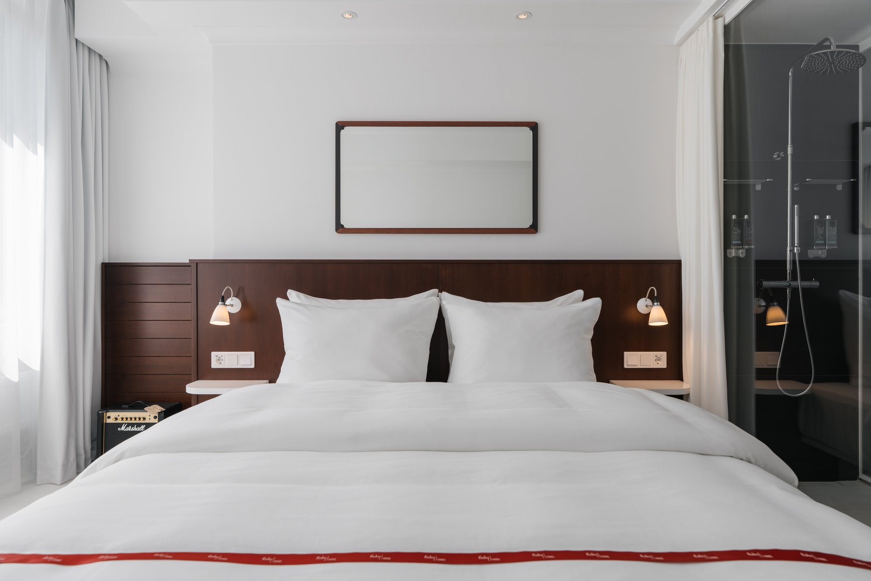 Doppelbett im Ruby Frida in Stockholm mit dunklem Holzkopfteil und weißer Bettwäsche mit rotem Zierband. Links am Bett steht ein kleiner Marshall-Verstärker, rechts grenzt die verglaste Dusche an.