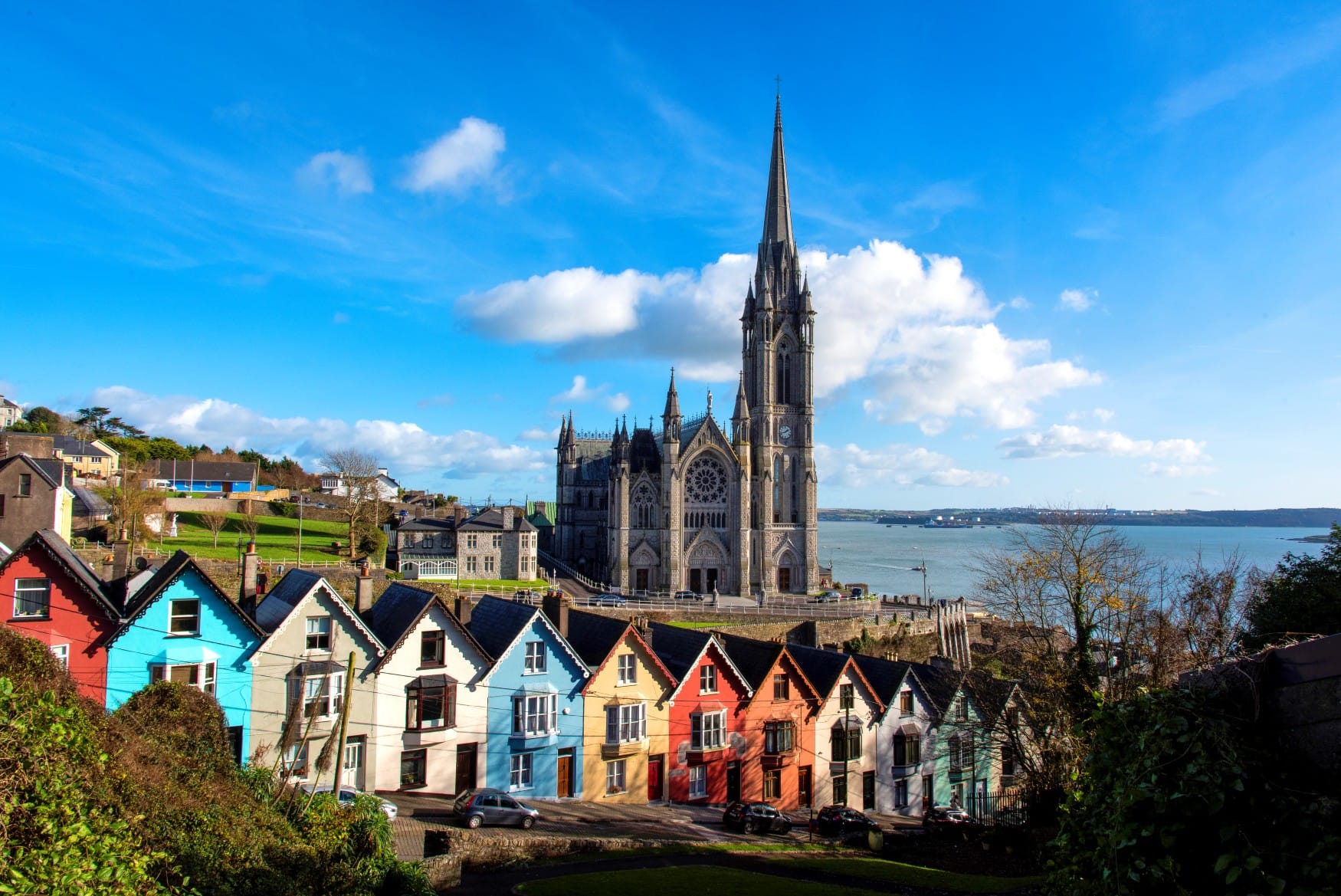 Blick über Cobh in der Grafschaft Cork mit der hoch aufragenden St Colman's Cathedral und einer Reihe bunter Reihenhäuser. Dahinter liegt der weite Naturhafen unter einem blauen Himmel.