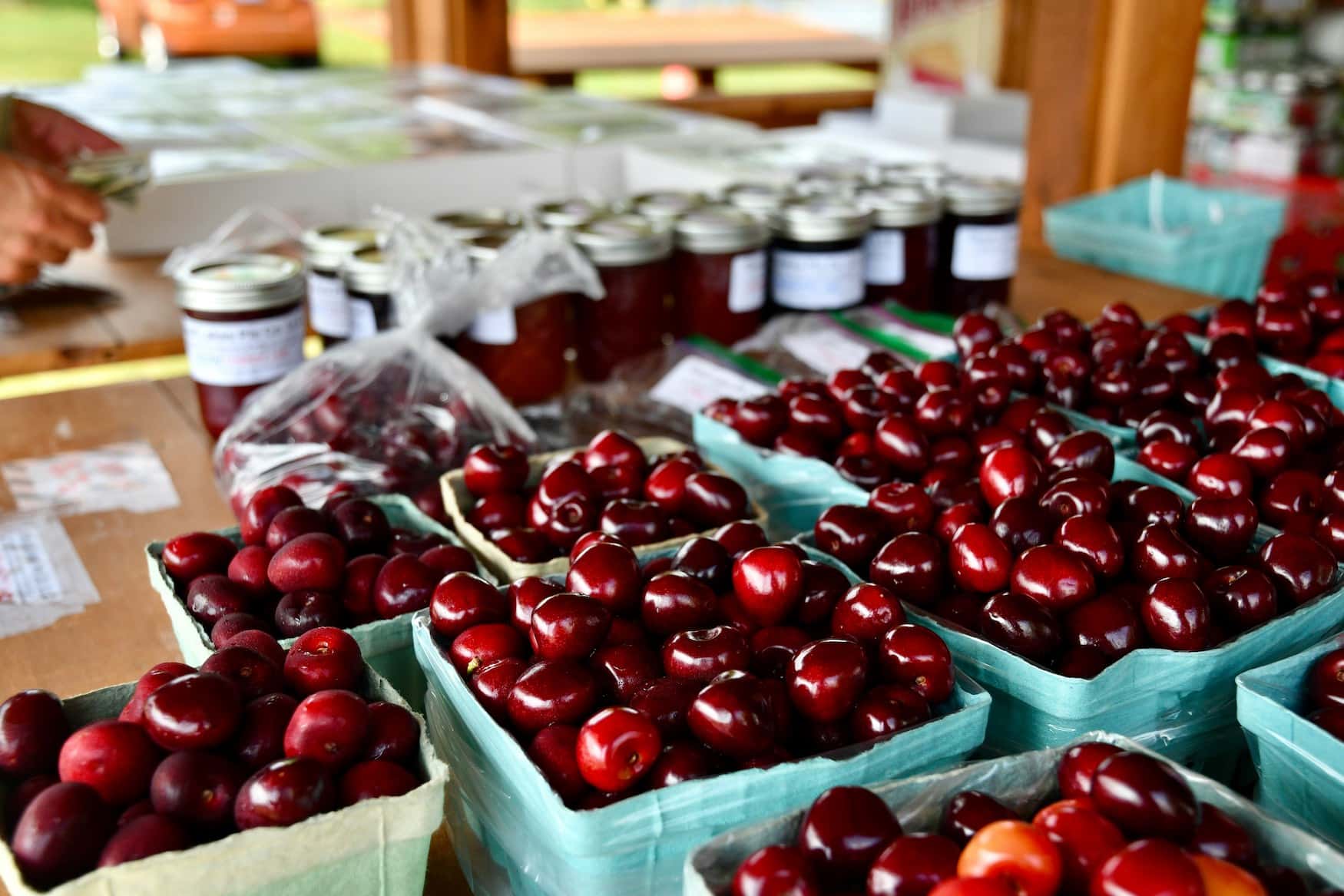 Frische rote Kirschen in Körbchen an einem Marktstand, dahinter Gläser mit eingekochten Früchten