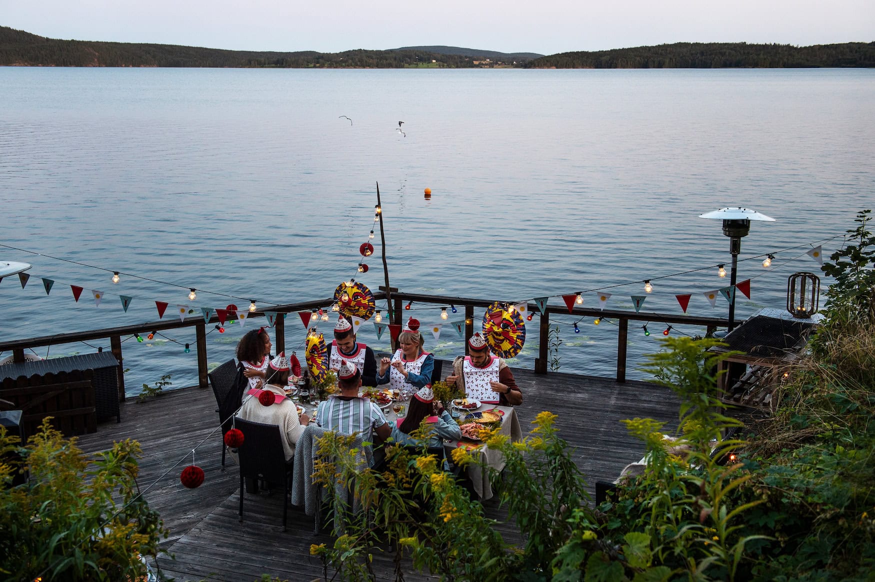 Gruppe feiert eine Kräftskiva an einer langen Tafel am See, mit Lampions, Girlanden und Papierhüten