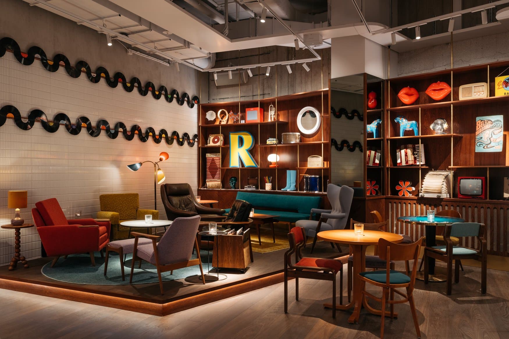 Lounge mit erhöhter Bühne im Ruby Frida in Stockholm. An der Wand sind Schallplatten zu Wellenlinien gehängt, in den Holzregalen stehen Dekorationsobjekte wie rote Lippen und blaue Dalapferde, davor Vintage-Sessel.