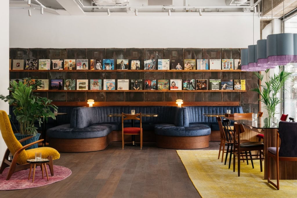 Loungebereich im Ruby Frida in Stockholm mit einer geschwungenen blauen Samtbank vor einer Wand aus Schallplattencovern. Links steht ein gelber Ohrensessel, rechts sind Tische mit Holzstühlen gedeckt.