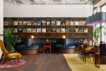 Loungebereich im Ruby Frida in Stockholm mit einer geschwungenen blauen Samtbank vor einer Wand aus Schallplattencovern. Links steht ein gelber Ohrensessel, rechts sind Tische mit Holzstühlen gedeckt.