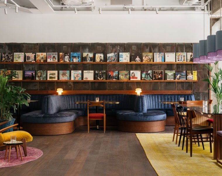 Loungebereich im Ruby Frida in Stockholm mit einer geschwungenen blauen Samtbank vor einer Wand aus Schallplattencovern. Links steht ein gelber Ohrensessel, rechts sind Tische mit Holzstühlen gedeckt.