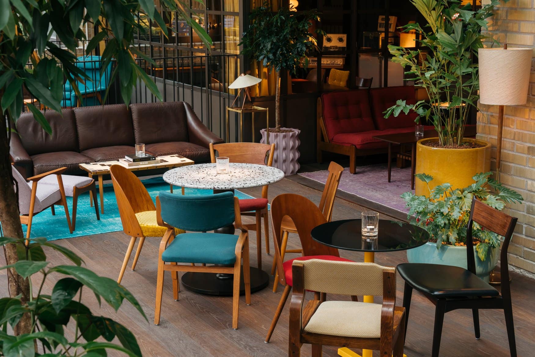 Loungebereich im Ruby Frida in Stockholm mit Sitzmöbeln im Stil der Mitte des 20. Jahrhunderts. Um einen runden Terrazzotisch stehen bunt gepolsterte Holzstühle, dahinter ein Ledersofa und viele Grünpflanzen.
