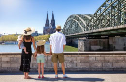 Eine junge Familie mit Kleinkind und Baby blickt vom Rheinufer auf dem Kölner Dom. Alle haben Strohhüte auf.