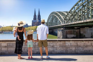 Eine junge Familie mit Kleinkind und Baby blickt vom Rheinufer auf dem Kölner Dom. Alle haben Strohhüte auf.