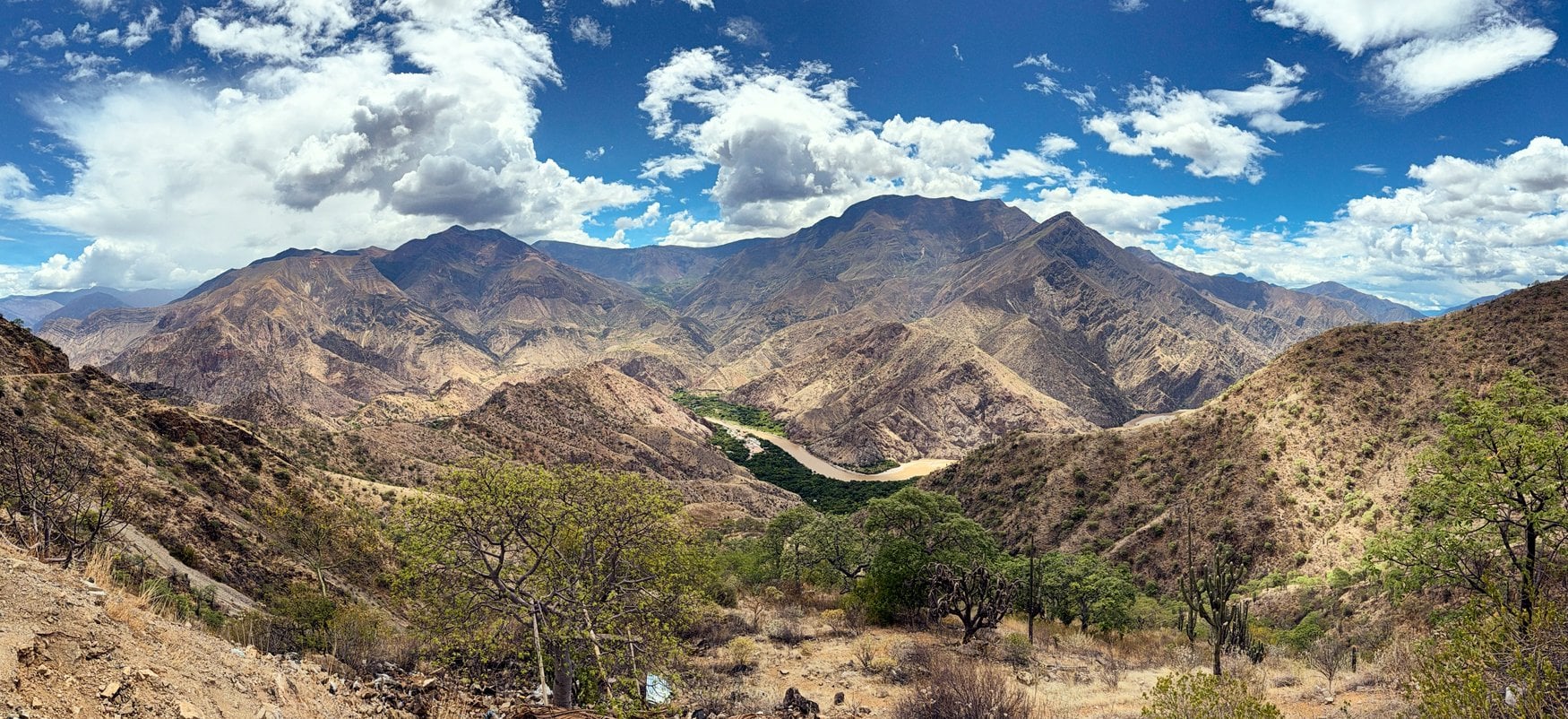 Panorama einer trockenen Andenlandschaft mit tief eingeschnittenem Flusstal. Über den kargen Bergrücken türmen sich weiße Wolken vor blauem Himmel.