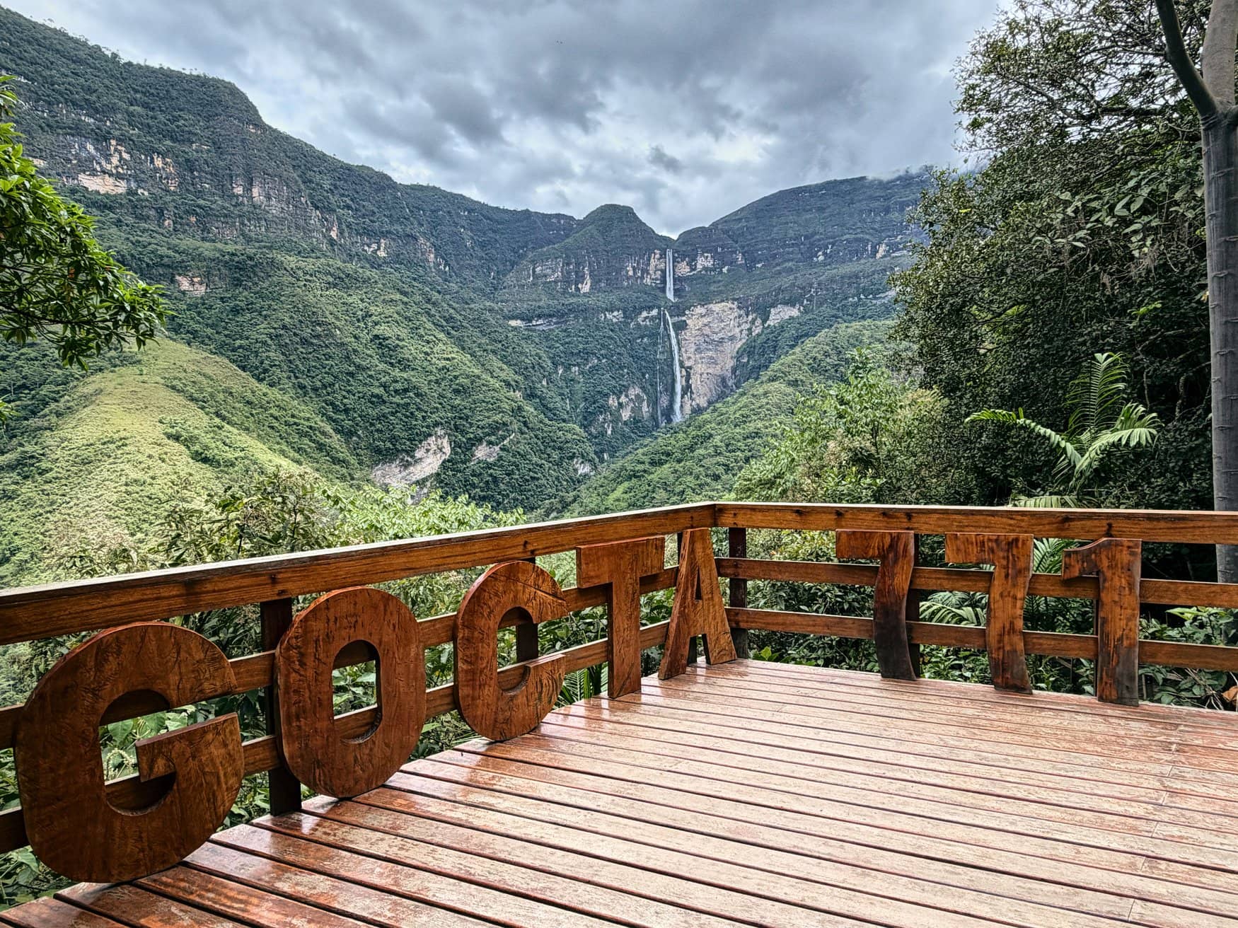 Holzterrasse eines Aussichtspunkts mit dem geschnitzten Schriftzug Gocta 771. Im Hintergrund stürzt der Gocta-Wasserfall über eine bewaldete Bergwand ins Tal.