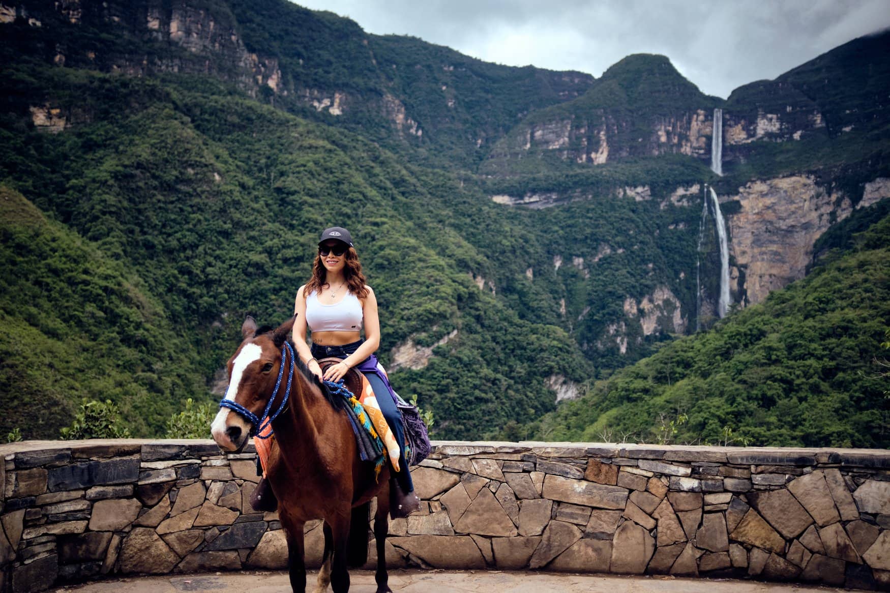 Eine Frau sitzt auf einem Pferd an einer Natursteinmauer eines Aussichtspunkts. Hinter ihr fällt der Gocta-Wasserfall die grün bewachsenen Anden hinab.