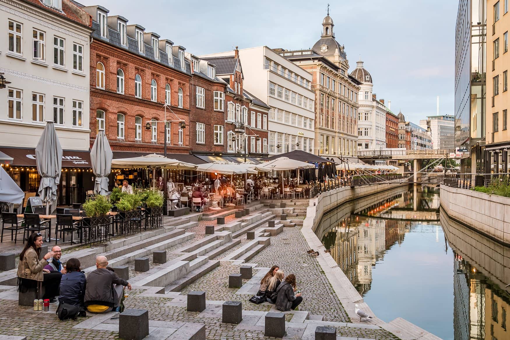 Menschen sitzen am Abend auf den Uferstufen des Aarhus å in Dänemark, entlang der Wasserkante reihen sich Cafés mit Sonnenschirmen vor historischen Häuserfassaden.