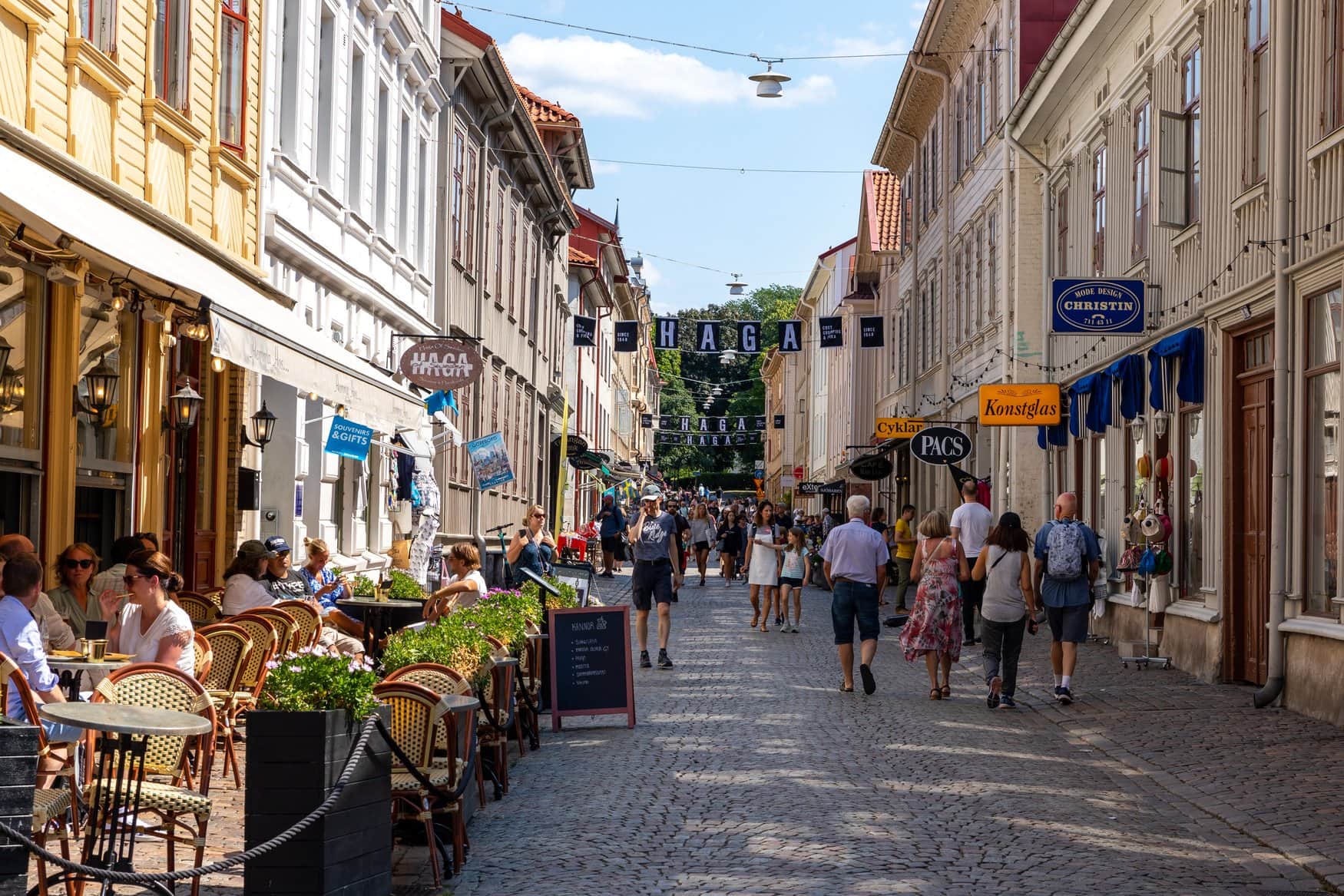 Belebte Kopfsteinpflastergasse im Viertel Haga in Göteborg mit Straßencafés und Läden entlang der Häuserzeilen und mehreren Schildern über der Gasse, darunter der Schriftzug Haga, bei sonnigem Wetter.