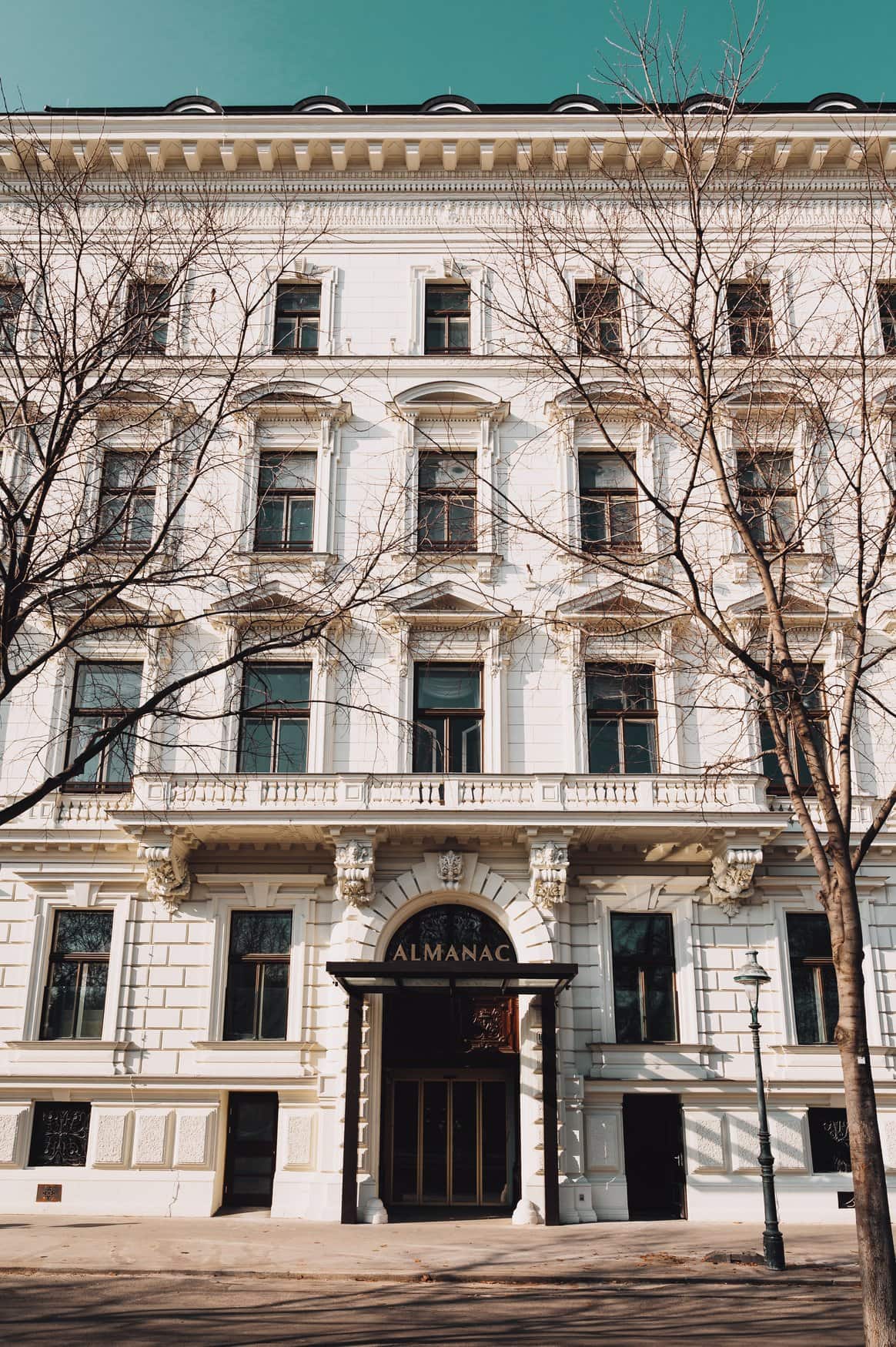 Historische Fassade des Almanac Palais Vienna mit hellem Stuck und dem Schriftzug Almanac über dem Eingang, davor kahle Bäume vor türkisfarbenem Himmel.