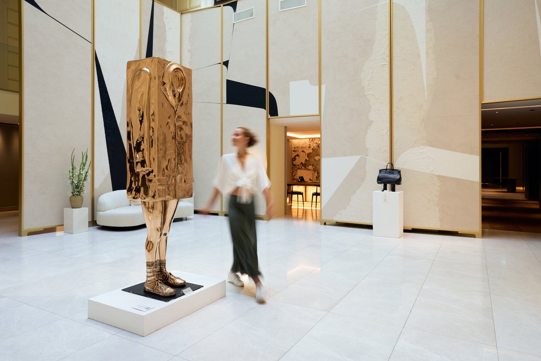 Goldglänzende Bronzeskulptur von Erwin Wurm in Gestalt eines Mantels auf zwei Beinen in der Lobby, im Hintergrund geht eine Frau in Bewegungsunschärfe vorbei.