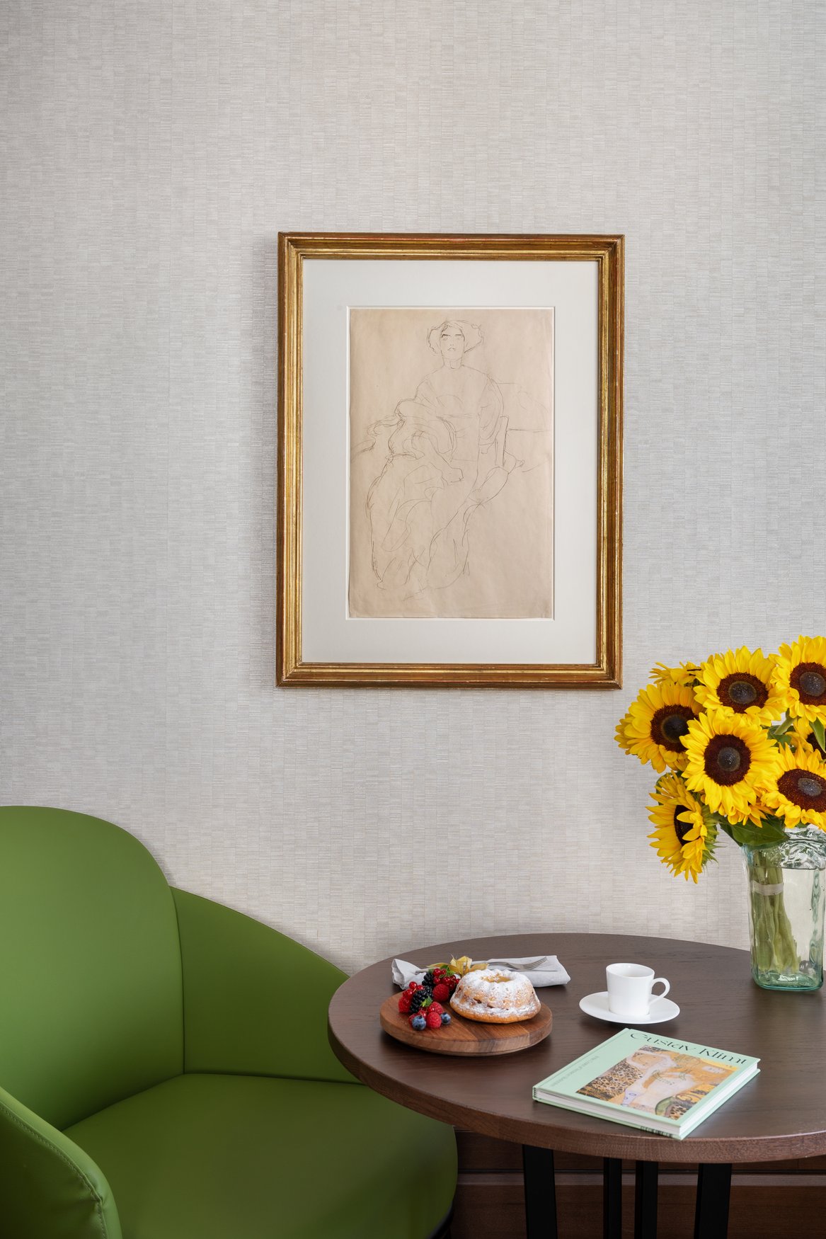 Wohnbereich der Klimt-Suite mit gerahmter Bleistiftzeichnung an der Wand und rundem Holztisch, auf dem ein Gugelhupf mit frischen Beeren und eine Espressotasse stehen, dazu gelbe Sonnenblumen und ein grüner Sessel.