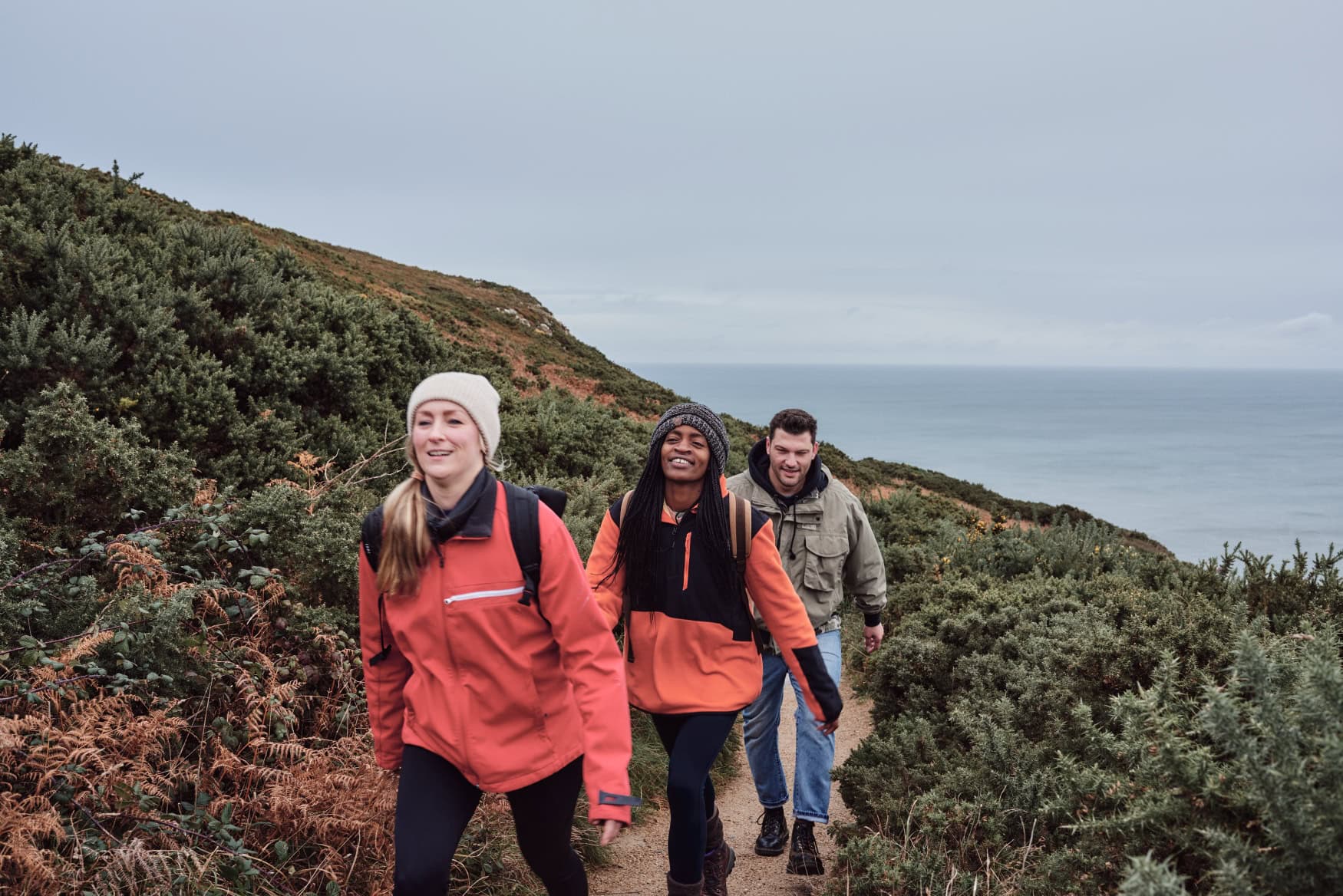 Drei warm gekleidete Personen wandern lächelnd auf einem Küstenpfad bei Howth nahe Dublin. Neben dem Weg wächst dichtes Grün, im Hintergrund liegt das graue Meer. 