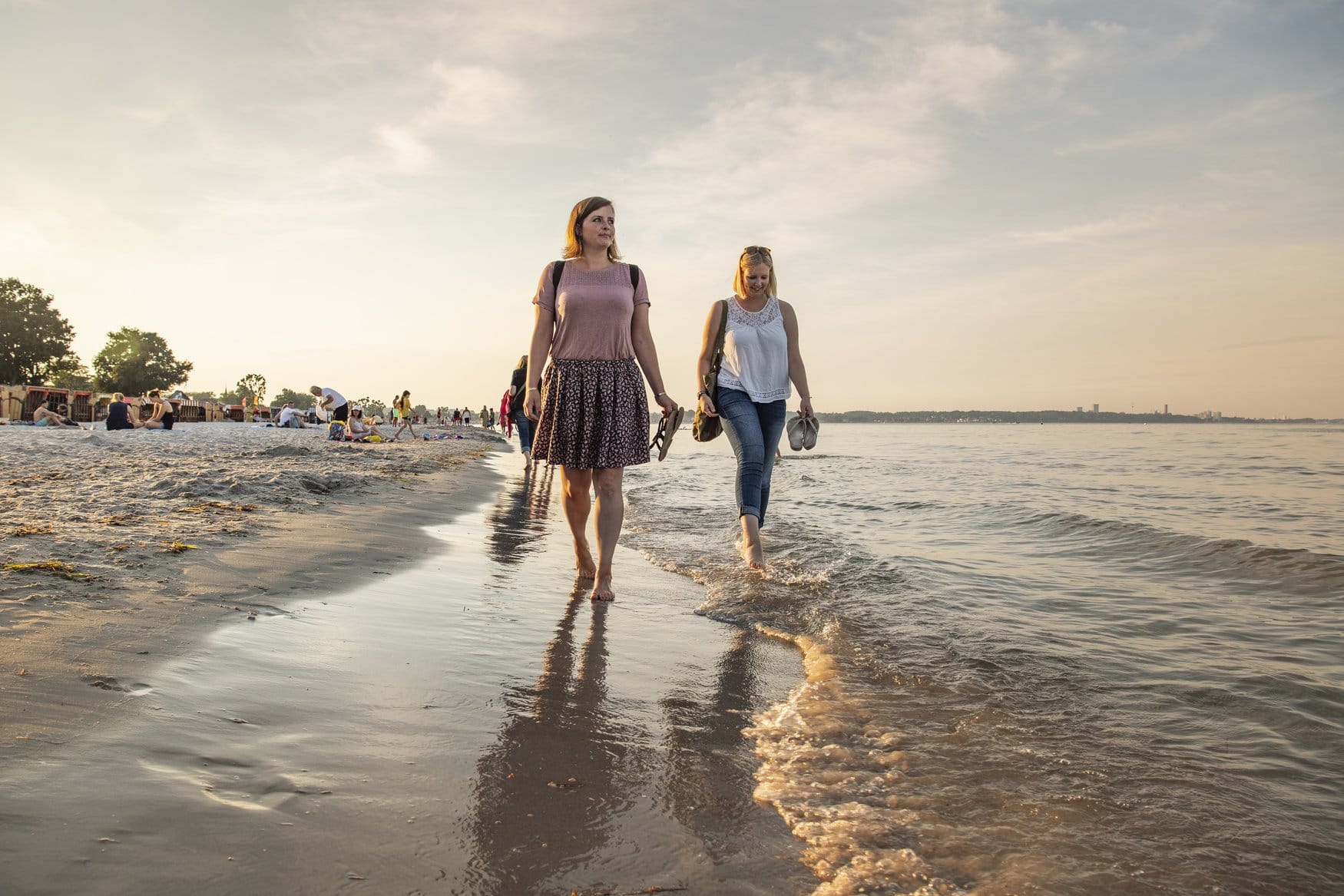 Zwei Frauen gehen barfuß am Wassersaum des Strandes von Scharbeutz entlang und tragen ihre Schuhe in der Hand. Im Hintergrund sind weitere Strandgäste zu sehen.