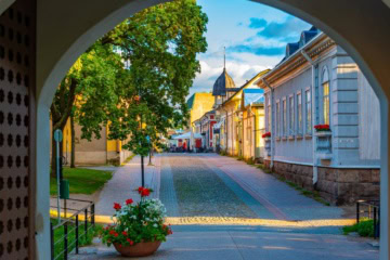 Blick durch einen steinernen Torbogen auf eine Kopfsteinpflastergasse in der Altstadt von Rauma mit bunten Holzhäusern, im Vordergrund steht ein Blumenkübel.