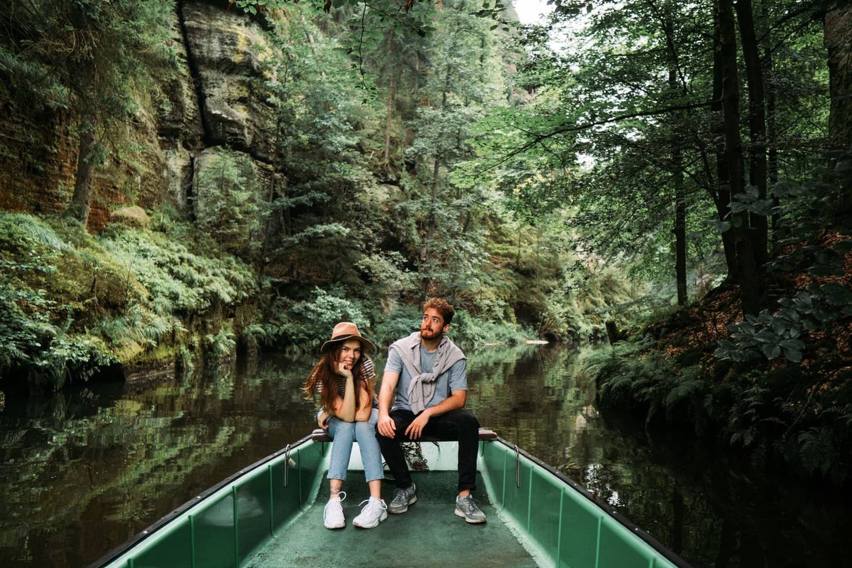 Ein Paar sitzt am Bug eines Ausflugsboots auf einer ruhigen Flussklamm in der Böhmischen Schweiz, umgeben von bewaldeten Felswänden.