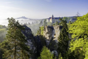 Blick über die Felsformationen von Groß-Skal im Böhmischen Paradies auf ein gelbes Schloss und die Ruine der Burg Trosky in der Ferne, morgens im Nebel.