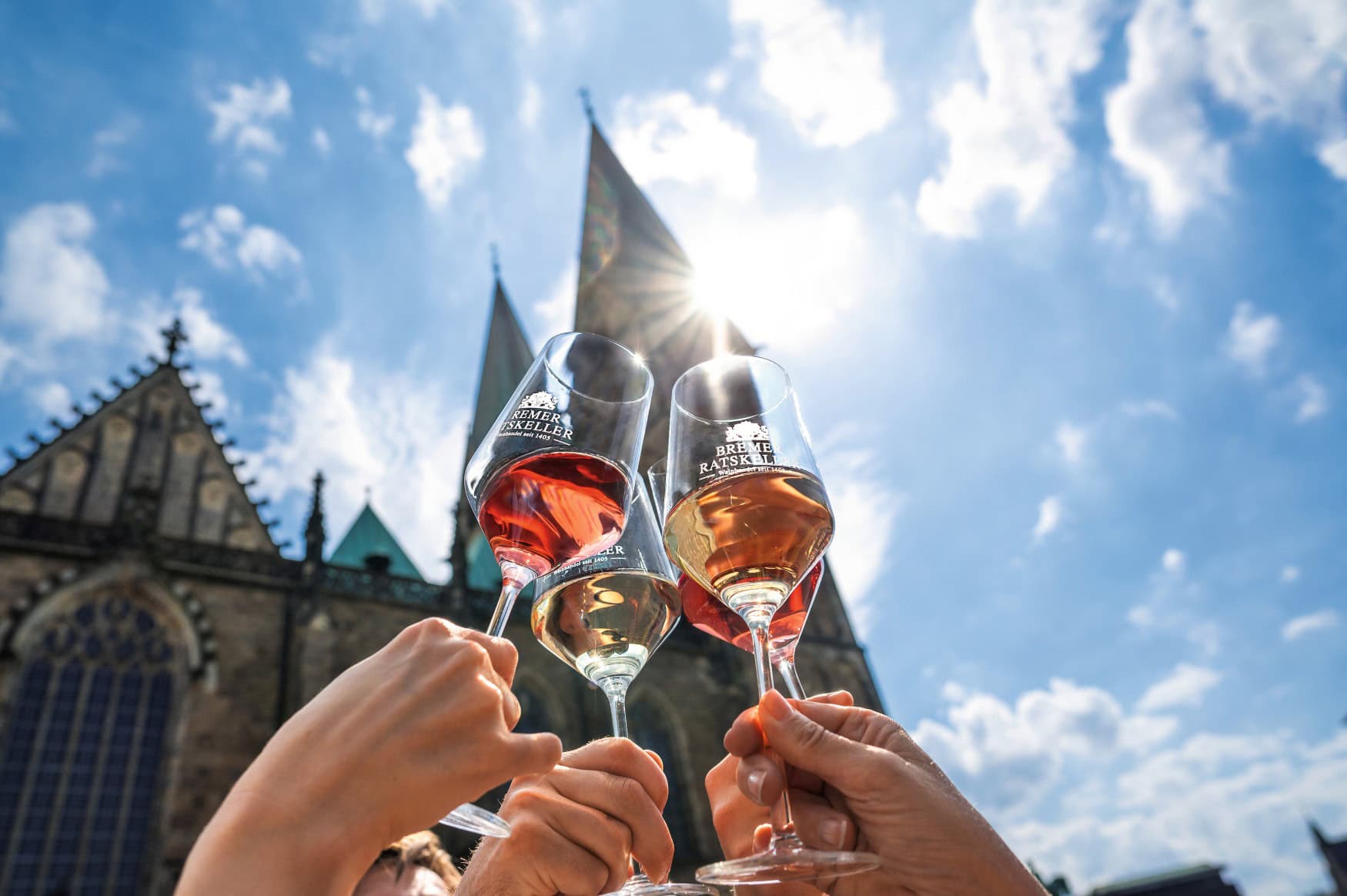 Drei Personen halten drei Gläser mit Rosé in die Höhe, im Hintergrund das Bremer Rathaus und blauer Himmel