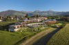 Drohnenaufnahme des Hotels Das Mühlwald in Südtirol, ein flach gebautes, moderens Resort in einer grünen Landschaft, umgeben von Weinstöcken. Im Hintergrund sind die Alpen sichtbar