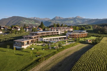 Drohnenaufnahme des Hotels Das Mühlwald in Südtirol, ein flach gebautes, moderens Resort in einer grünen Landschaft, umgeben von Weinstöcken. Im Hintergrund sind die Alpen sichtbar