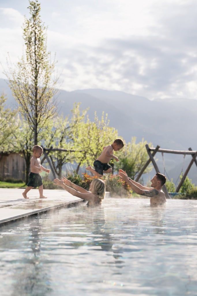 Ein Mann und eine Frau stehen im Wasser des Outdoor-Pools eines Hotels, die Arme jeweils zum Beckenrand gestreckt. Von dort rennen zwei Kleinkinder freudig in Richtung der Eltern.