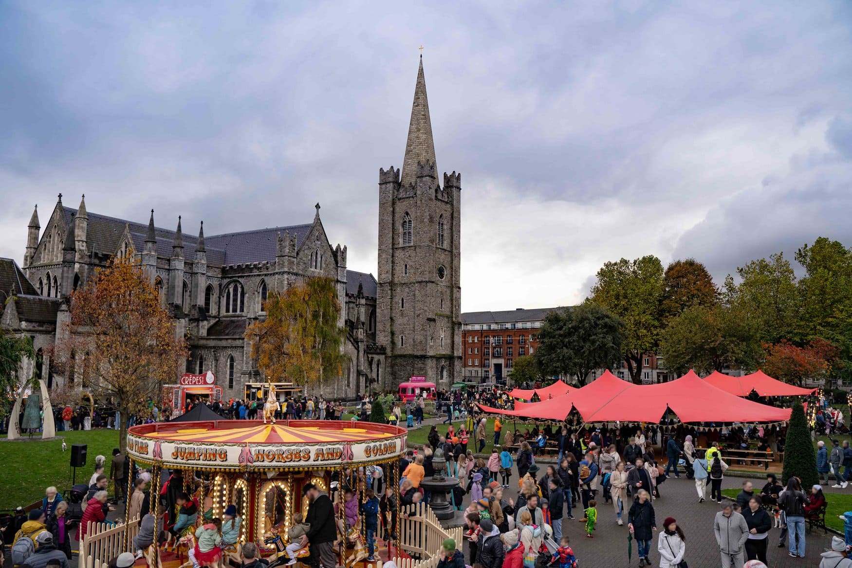 Das Bram Stoker Festival im Park an der St Patrick's Cathedral in Dublin. Auf der Wiese dreht sich ein historisches Karussell, rechts stehen rote Zeltdächer über Bierbänken, dazwischen bewegen sich Besucher.