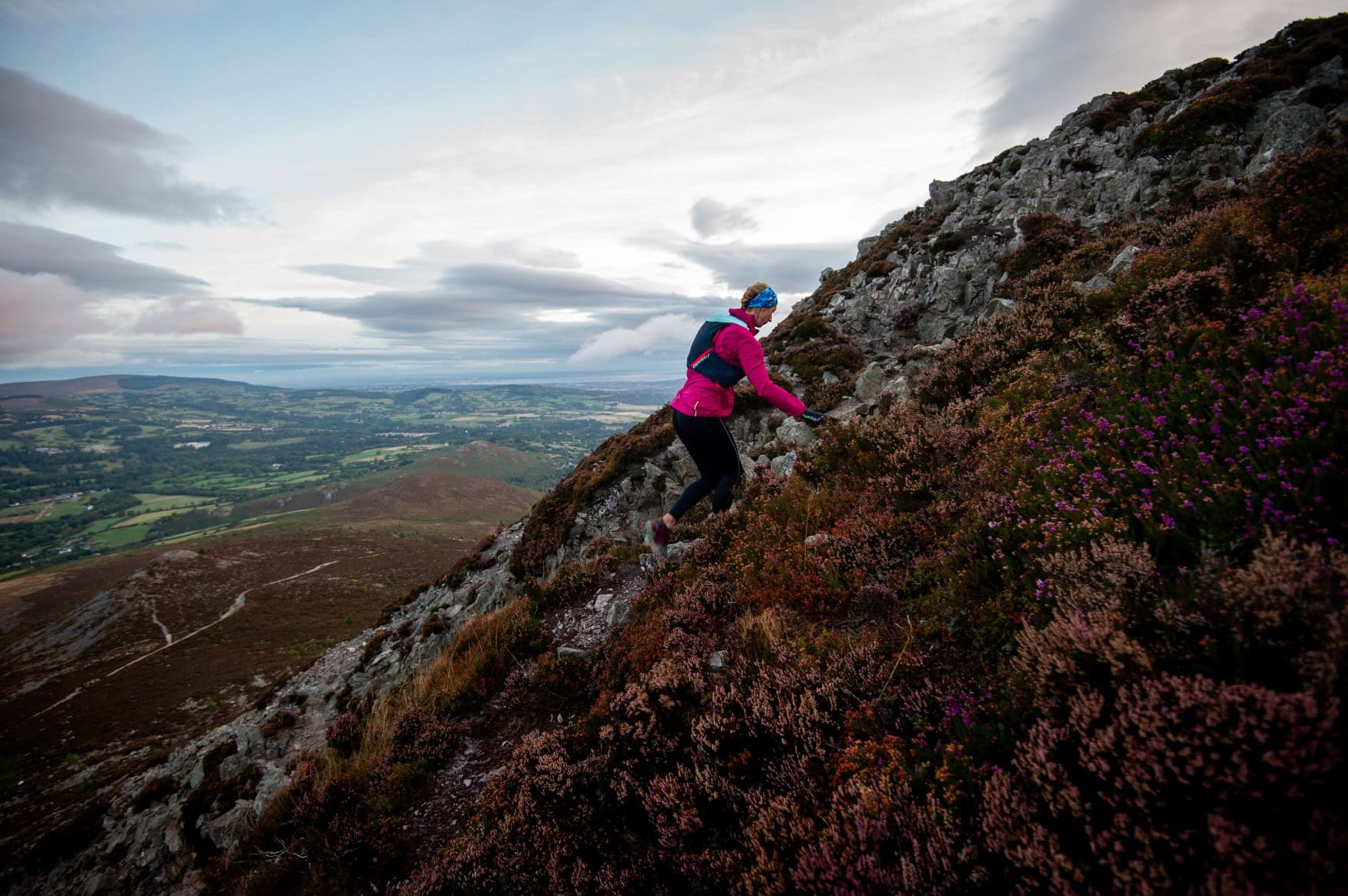 Eine Frau in Wanderkleidung steigt einen felsigen Wanderweg in Irland hinauf, im Hintergrund eine weite Landschaft.