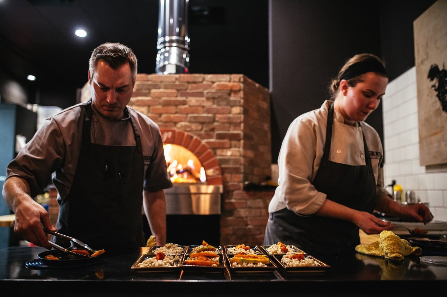 Zwei Köche richten Vorspeisen auf schwarzen Tellern an, im Hintergrund der brennende Holzofen der Restaurantküche