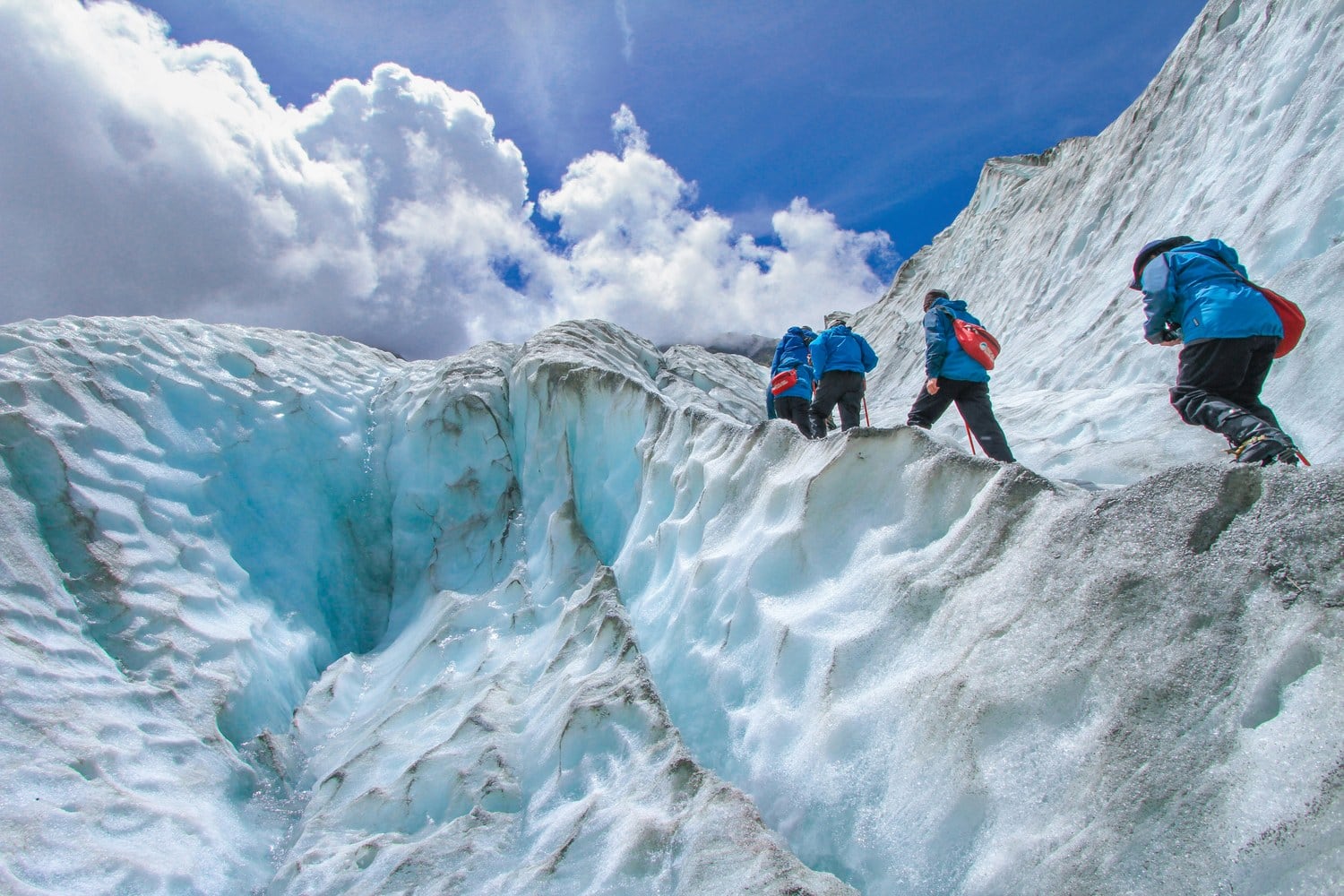 Vier Eiswanderer in blauen Jacken balancieren über einen Grat zwischen zwei tiefen Gletscherspalten