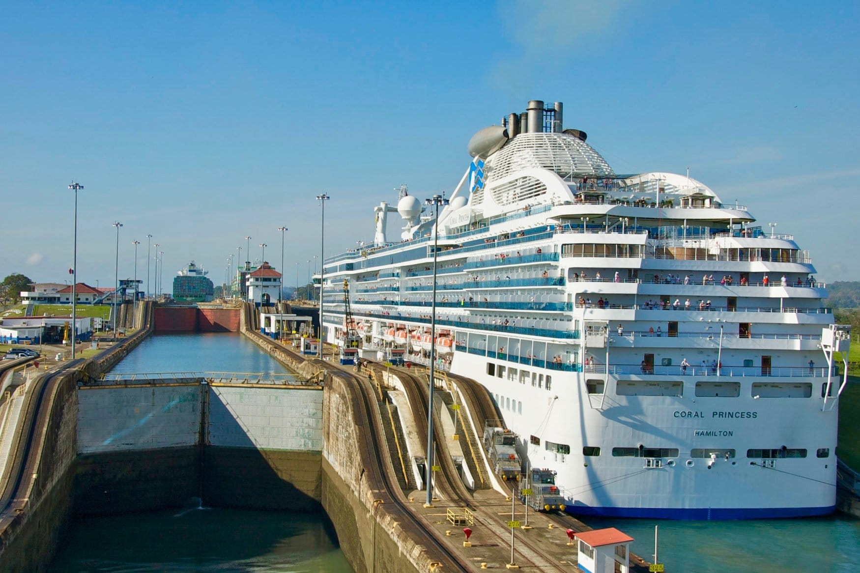 Das Kreuzfahrtschiff Coral Princess liegt in einer Kammer der Gatún-Schleusen am Panamakanal. Passagiere stehen an den Decksreihen, neben dem Rumpf verlaufen die Schienen der Treidellokomotiven.