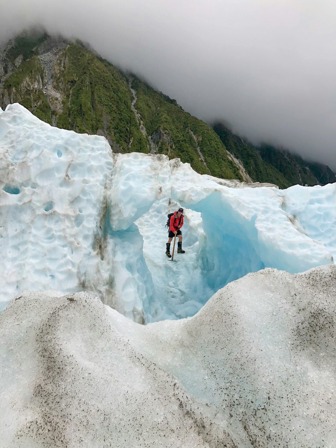 Einzelner Wanderer in roter Jacke steht in einer schmalen Eisspalte, dahinter ein nebelverhangener Regenwaldhang