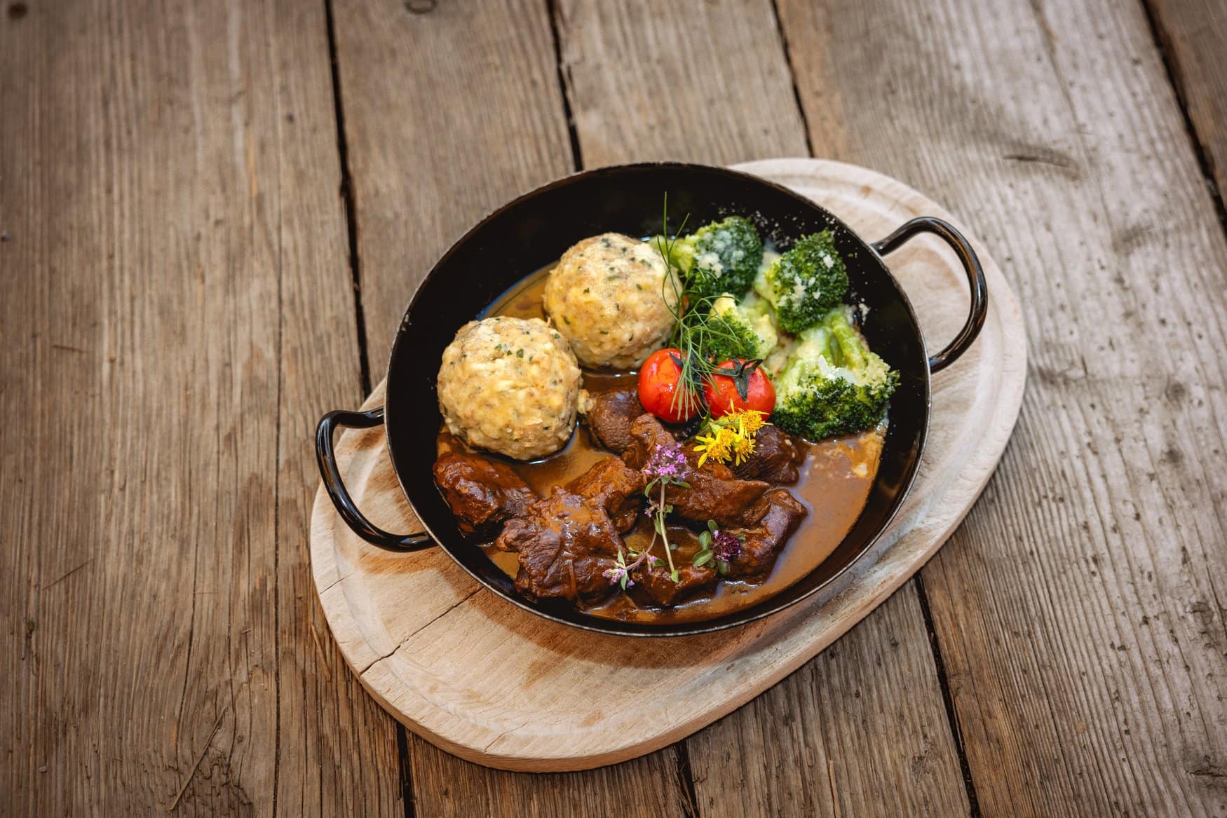 Gulasch mit zwei Knödeln, Broccoli und Kirschtomaten in einer schwarzen Pfanne auf einem Holzbrett.