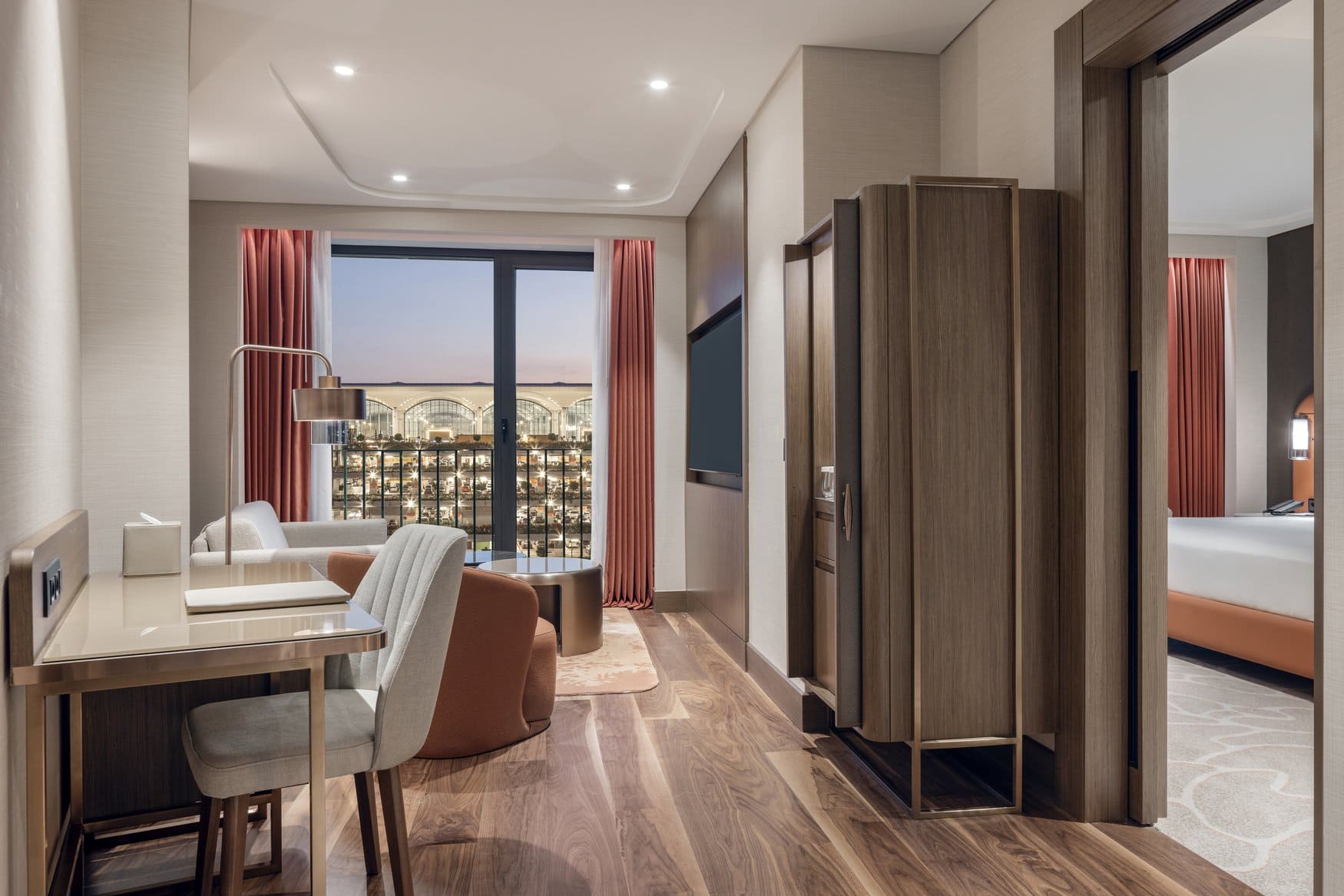 Wohnbereich einer Suite im Hilton Istanbul Airport mit Schreibtisch, Sesseln und Fernseher. Durch die Fensterfront fällt der Blick bei Dämmerung auf das Terminaldach des Flughafens, rechts öffnet sich der Durchgang zum Schlafzimmer.